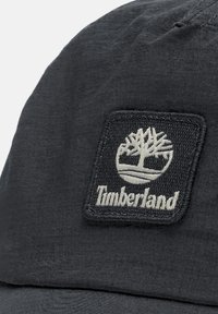 Cappello nero realizzato in tessuto testurizzato, con un patch quadrato ricamato raffigurante un logo ad albero e la scritta "Timberland" in bianco.