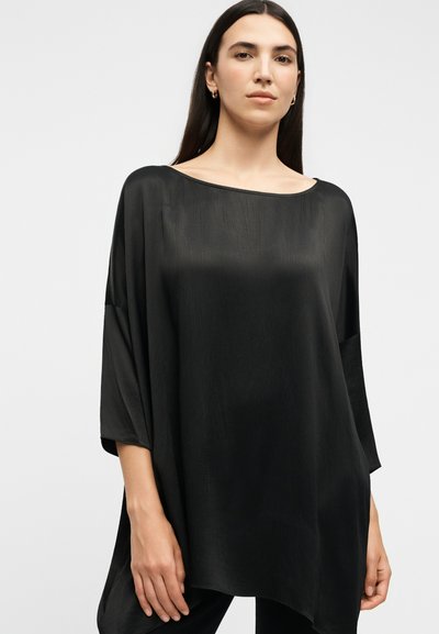 Žena s dugom tamnom kosom nosi lepršavu crnu satensku bluzu široka brodskog izreza i rukava do pola podlaktice na jednobojnoj pozadini.
