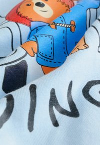 Bavlnená tkanina s farebnou ilustráciou medveďa v modrom kabáte, obklopená čiernym textom na svetlomodrom pozadí.