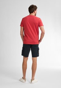 Camiseta de algodón roja, de mangas cortas, con cuello redondo, combinada con pantalones cortos cargo negros, que cuentan con bolsillos laterales y zapatillas con acentos en color beige.