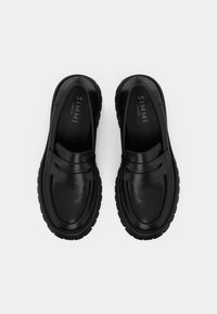 Čierne kožené mokasíny s hladkým povrchom a hrubou gumovou podrážkou. Majú dekoratívny remienok na prednej strane. Vnútorné označenie značky je viditeľné.