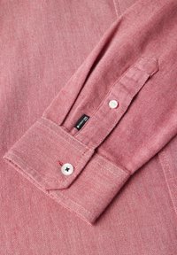 Camicia rossa con texture, dotata di polsini con bottoni, caratterizzata da bottoni bianchi e un'etichetta nera. Il tessuto presenta un sottile motivo a spina di pesce.