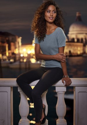 Young woman with curly hair wearing a gray top and black pants sits on a stone railing with illuminated city buildings in the background at night.