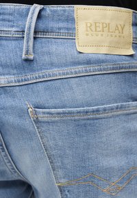Κοντινή λήψη ανοιχτό μπλε τζιν παντελονιού που εμφανίζει την πίσω τσέπη, την θηλιά ζώνης και το μπεζ δερμάτινο ταμπελάκι με την επωνυμία "Replay Blue Jeans".