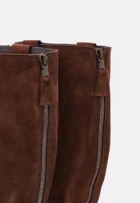 Botas de tobillo de ante marrón con cremalleras plateadas a ambos lados y una textura suave. Presentan un borde cosido y un diseño minimalista.