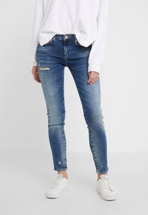 Niebieskie dżinsy o wąskim kroju z lekkim przetarciem, noszone z białą bluzką z długim rękawem i białymi trampkami na jednolitym tle.