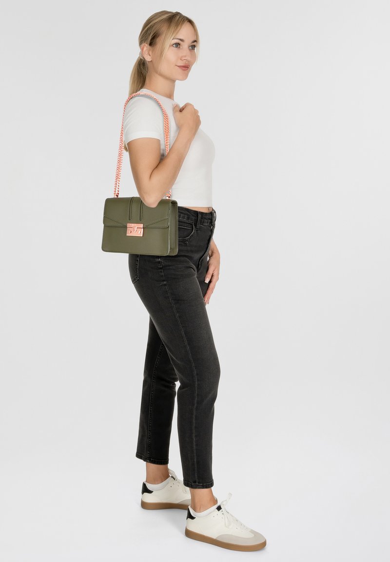 Bolso de mano verde olivo con un diseño estructurado, herrajes en oro rosa y una correa trenzada. La modelo lleva una blusa blanca ajustada y jeans negros.