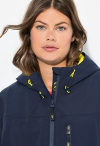 Veste bleu marine avec fermeture éclair à l'avant, capuche avec accents jaunes et cordons de serrage réglables. Dotée d'un tissu lisse et texturé, ainsi que d'une poche avant.