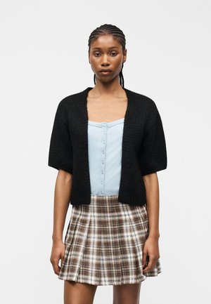 Vrouw met gevlochten haar, gekleed in een zwart kort mouw vest over een lichtblauw knoopjes shirt en een geruite bruine geplooide rok, staand.