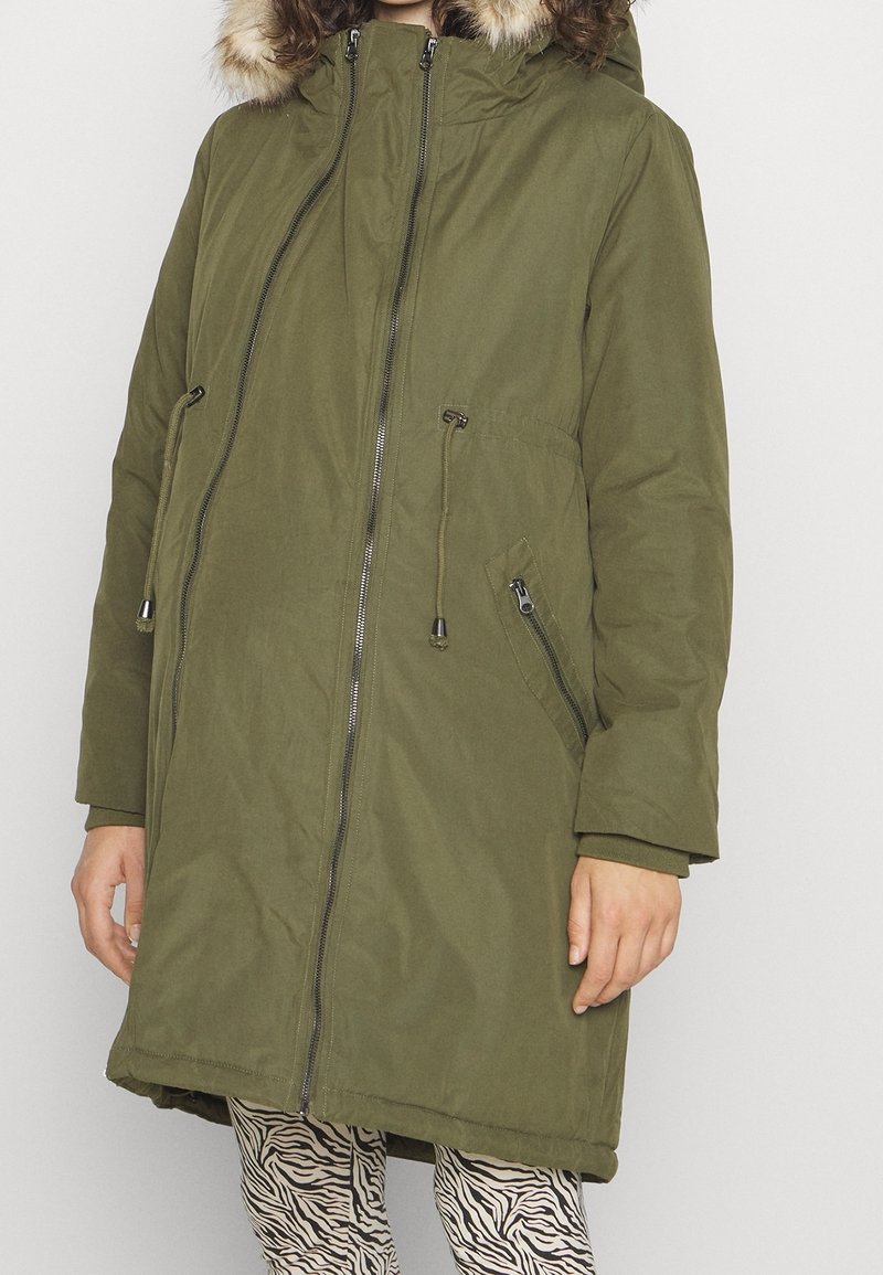 Cazadora tipo parka verde oliva con capucha forrada de piel sintética, dos bolsillos laterales con cremallera y cierre de cremallera. Llevada sobre leggings estampados.