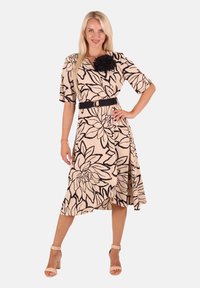 Beige Midi-Kleid mit schwarzem Blumenmuster, V-Ausschnitt, kurzen Ärmeln, tailliertem Bund mit schwarzem Gürtel und ausgestelltem Saum. Getragen mit beigen Absätzen.