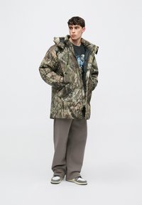 Giacca a vento imbottita mimetica con cappuccio, caratterizzata da un motivo testurizzato simile a corteccia nei toni del verde e marrone. Indossata sopra una t-shirt nera con stampa grafica e pantaloni grigi.