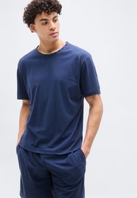 Άνδρας με σγουρά μαλλιά που φοράει μπλε σκούρο κοντομάνικο πουκάμισο και ταιριαστά σορτς, στέκεται με τα χέρια στις τσέπες, κοιτάζοντας προς το πλάι.