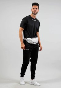 Uomo in abbigliamento sportivo Givova nero e sneakers bianche sta in piedi indossando una marsupio Givova bianca su uno sfondo grigio uniforme.