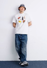 White cotton t-shirt featuring various animal graphics, paired with blue denim jeans and a checkered bucket hat. Grey athletic sneakers.