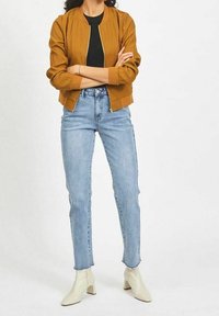 Hořčicová bomber bunda se zipem vpředu, v kombinaci s světle modrými skinny džíny a bílými kotníkovými botami. Hladká tkanina a přiléhavý střih.