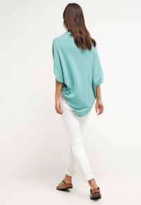 Ljus teal ponchosweater i mjukt tyg, med en lös passform, fladdermusärmar och rundad bakre fåll, kombinerad med vita rullade jeans och sandaler.