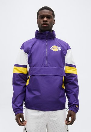 Άνδρας που φοράει μοβ μπουφάν Los Angeles Lakers με κίτρινες και λευκές ρίγες, στέκεται μπροστά σε απλό ανοιχτόχρωμο φόντο.