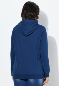 Osoba oblečená v jednoduché tmavě modré mikině a modrých džínech, zobrazená zezadu na bílém pozadí.