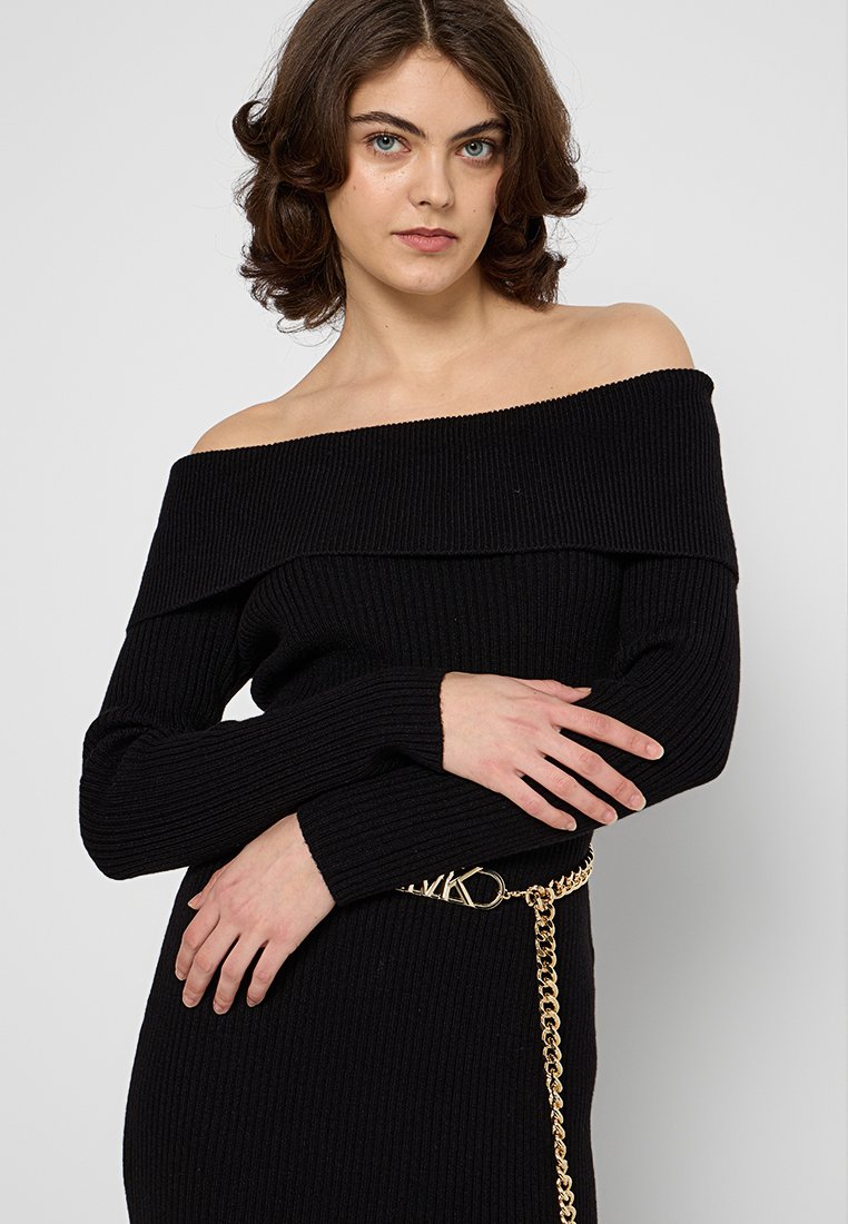 Abito nero a spalle scoperte in maglia a costine con maniche lunghe, caratterizzato da un dettaglio con cintura in catena dorata e silhouette aderente.