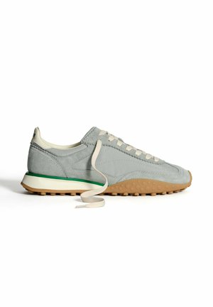 Zapatilla de ante gris con una suela texturizada, cordones blancos y un acento verde en la entresuela. Presenta un perfil bajo y un diseño de punta redonda.