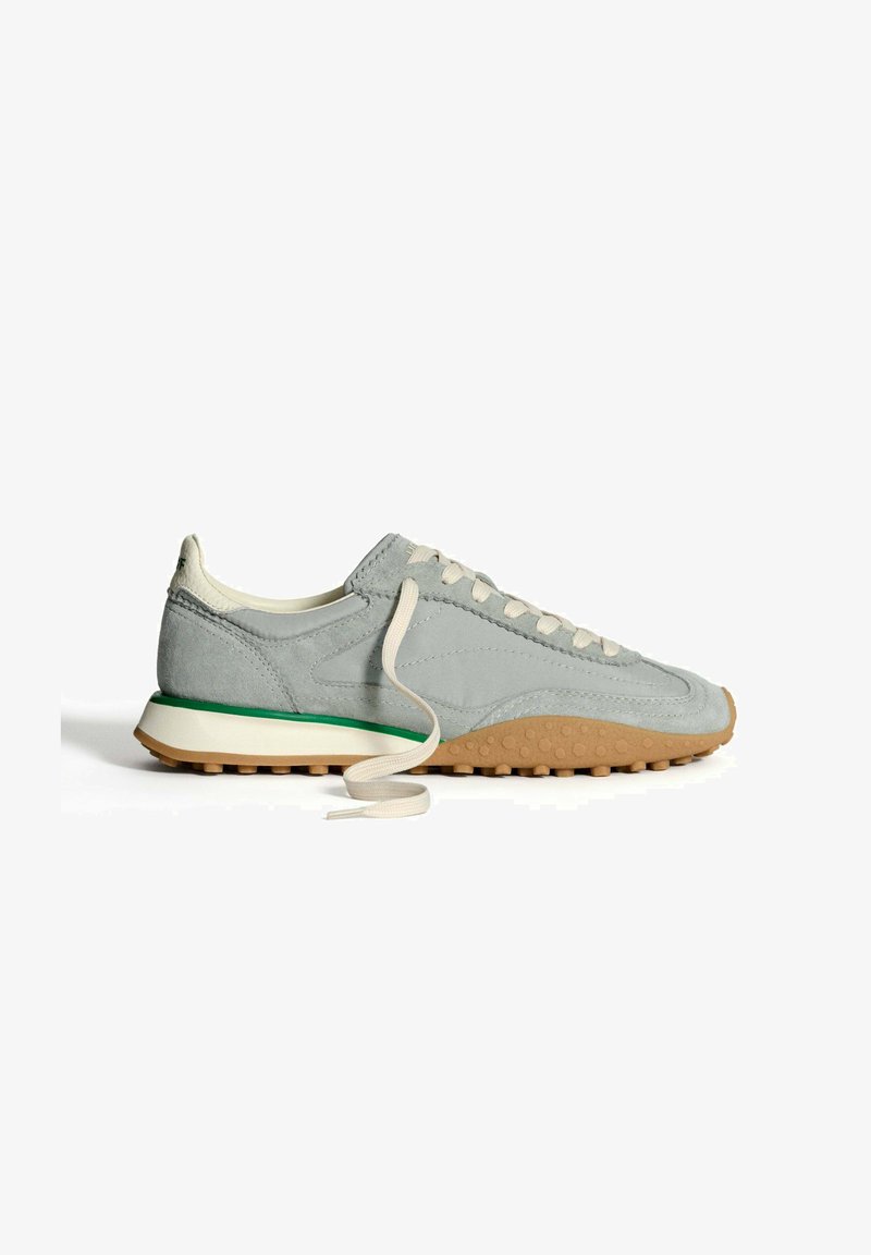 Zapatilla de ante gris con una suela texturizada, cordones blancos y un acento verde en la entresuela. Presenta un perfil bajo y un diseño de punta redonda.