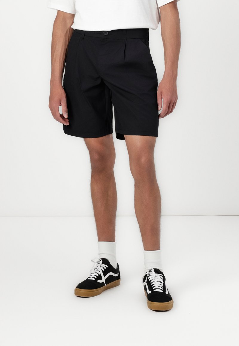 Shorts noirs en tissu léger avec un détail de pli sur le devant, associés à des baskets noires et des chaussettes blanches mi-mollet.