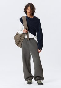 Marineblauer Oversized-Pullover über einem weißen Langarmshirt, graue weite Sweatpants und grüne Schuhe. Das Modell hält eine braune Tasche.
