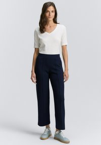 Maglietta grigia chiara a scollo a V, maniche corte; pantaloni larghi blu con cuciture frontali; sneakers blu con lacci bianchi e suola beige.