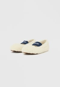 Par de zapatillas peludas color crema con bandas elásticas azul marino etiquetadas con "MOON BOOT" sobre un fondo blanco y liso.