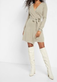 Nainen pukeutuneena beigeen ympärille kiedottuun mekkoon, jossa on sidottu vyö, ja valkoisiin korkeisiin nahkasaappaisiin, seisoo valkoista taustaa vasten.