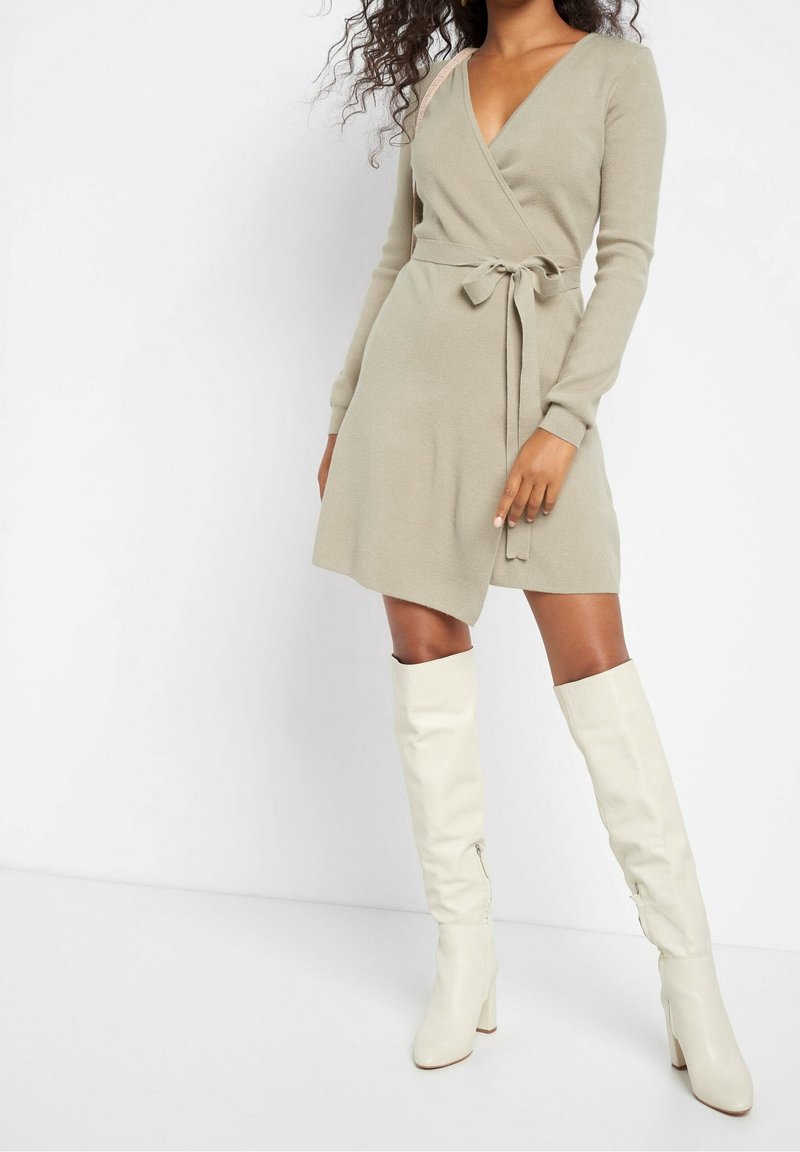 Nainen pukeutuneena beigeen ympärille kiedottuun mekkoon, jossa on sidottu vyö, ja valkoisiin korkeisiin nahkasaappaisiin, seisoo valkoista taustaa vasten.