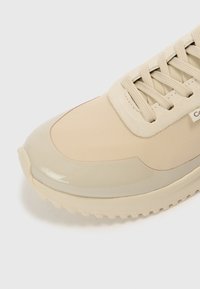 Beżowy sneaker z gładkim, błyszczącym czubkiem, teksturowanym zamszowym obszarem sznurowania i płaskimi sznurowadłami. Gumowa podeszwa z małymi wzorami na bieżniku.