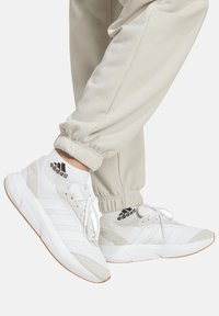 White athletic sneakers with mesh and suede panels, featuring three white stripes, laces, and a textured rubber sole. Paired with beige joggers.