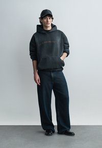 Tânăr care poartă o șapcă neagră, hanorac oversize întunecat, blugi largi de culoare închisă și pantofi negri, stând pe un fundal simplu, deschis la culoare.