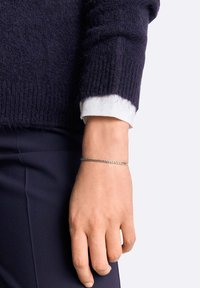Bracciale in argento su un polso, caratterizzato da un design elegante con maglie intrecciate, posizionato accanto a un maglione scuro e al polsino di una camicia bianca.