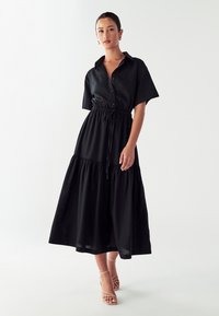 Juodas maxi suknios modelis su trumpomis rankovėmis, suveržtu liemeniu ir sluoksniuotu sijonu. Sklandžiai tekstūruota medžiaga, atsipalaidavęs siluetas ir švelnus blizgesys.