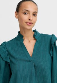 Blusa teal in tessuto texturizzato, con scollatura a V e dettagli arricciati sul colletto e sulle maniche. Presente un motivo a righe verticali.