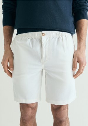 Hombre que lleva pantalones cortos blancos hasta la rodilla con cintura elástica y botón, combinados con una camiseta de manga larga azul oscuro.