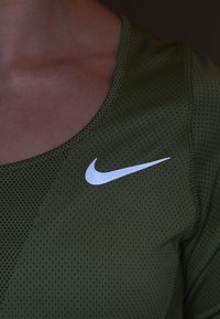 Olivegrünes Sporttop mit atmungsaktivem Mesh-Material, versehen mit einem weißen Nike Swoosh-Logo auf der linken Brust.