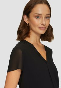 Musta pusero lyhyillä läpinäkyvillä hihoilla, V-pääntiellä ja poimutetulla designilla. Silkkinen kangas, jossa on hieno kiilto. Yksinkertainen ja elegantti.