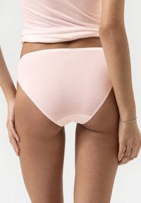Femme portant des culottes en coton rose clair et un haut assorti, vue de dos, se tenant contre un fond blanc uni.
