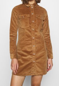 Brun cordfløyelsklänning med lång ärm, knappstängning fram, två bröstfickor och silverknappar. Mjuk textur och skräddarsydd passform.