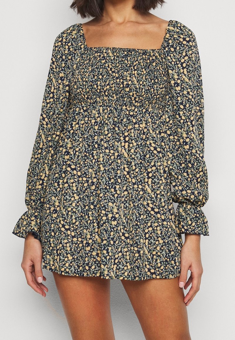 Femme portant une robe courte à fleurs à manches longues, avec un col carré et un corsage smocké dans des tons bleu foncé et jaune.