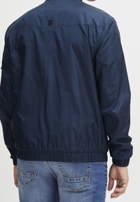 Marineblauwe nylon jas met een gladde textuur, elastische tailleband en boorden; heeft een klein logoaccent op de achterkant.