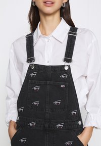 Svarta denimsoveraller med flera broderade Tommy-logotyper, silverfärgad metall, justerbara band och en framficka.