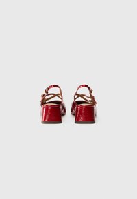 Chaussures en cuir verni rouge avec un petit talon carré, présentant un dos ouvert, des brides croisées et un détail de boucle. Surface texturée.