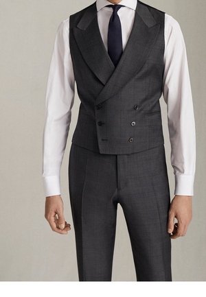Homme portant un gilet croisé gris foncé, pantalon assorti, chemise blanche et cravate noire sur fond uni.