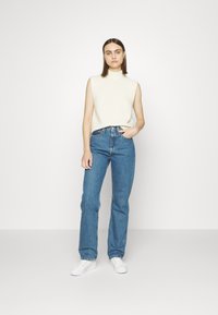 Λευκό χωρίς μανίκια πουλόβερ, ψηλό λαιμόκοψιμο, σε συνδυασμό με ανοιχτό μπλε παντελόνι φάρδου και λευκά αθλητικά παπούτσια; μινιμαλιστικός σχεδιασμός και άνετη εφαρμογή.