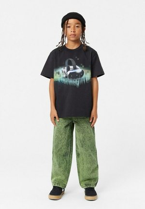 Svart bomullströja med en grafisk bild av en virtuell verklighetsheadset, kombinerad med vida gröna denimbyxor och svarta sneakers.